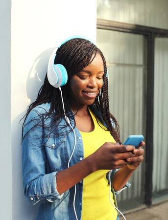 Beautiful smiling african woman with headphones listens to music and using smartphone in cityの写真素材