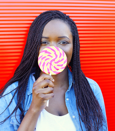 Portrait happy smiling african woman with sweet lollipop over red backgroundの写真素材