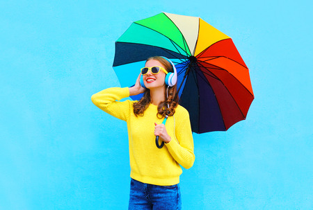 Lifestyle portrait smiling young woman listens to music in headphones with colorful umbrella in autumn day over colorful blue background wearing a yellow knitted sweaterの写真素材