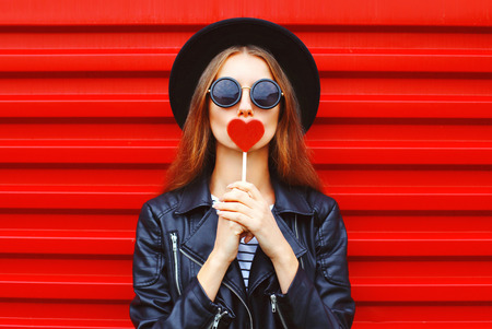 Fashion pretty young woman with red lollipop heart wearing black hat leather jacket over urban backgroundの写真素材