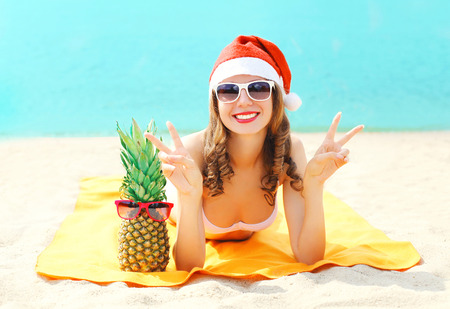 Christmas portrait pretty young smiling woman in red santa hat and pineapple lying on beach over sea...