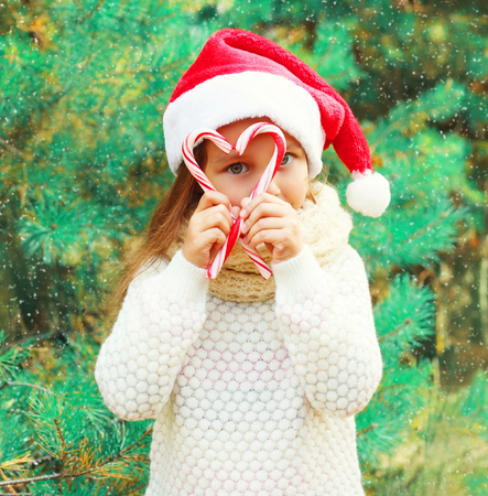 Christmas child little girl in santa red hat playing with sweet lollipop cane near a treeの写真素材