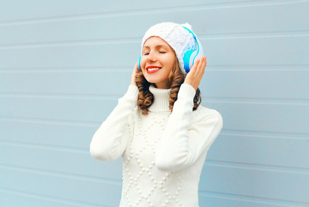 Happy young woman in headphones enjoys listens to music wearing a knitted hat, sweater over blue backgroundの写真素材