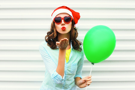 Portrait pretty woman in christmas red santa hat, with air balloon, heart sunglasses sends air kiss over white backgroundの写真素材
