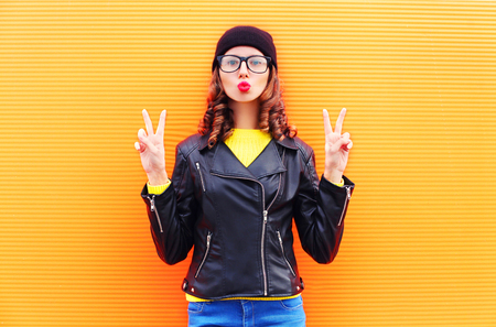 Fashion pretty young woman blowing red lips making air kiss wearing a black hat, rock jacket over colorful orange backgroundの写真素材