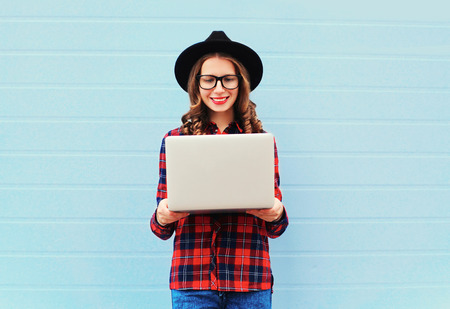 Fashion young smiling woman holding laptop computer in city, wearing black hat, red checkered shirt over blue backgroundの写真素材