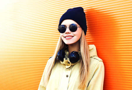 Fashion portrait happy blonde woman with headphones, sunglasses and black hat on a orange colorful background in cityの写真素材