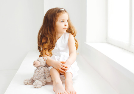 little girl child with teddy bear sitting at home in white room looks with hope in the windowの写真素材