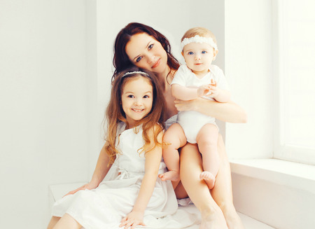 Happy mother with two her daughters children at home in white room sitting together near windowの写真素材