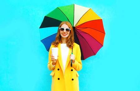 Happy smiling woman with coffee cup and colorful umbrella in yellow coat jacket on blue backgroundの写真素材