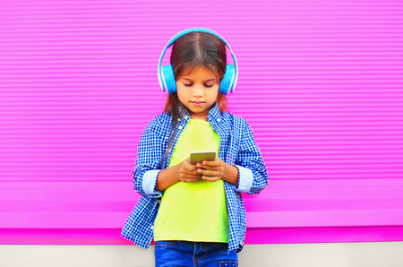 Little girl child with smartphone listens to music in wireless headphones on colorful pink wall backgroundの写真素材