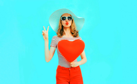 Beautiful young woman with red heart shaped balloons sending sweet air kiss on colorful blue backgroundの写真素材