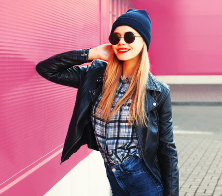 portrait stylish blonde smiling woman in rock black style jacket, hat posing on city street over colorful pink wall backgroundの写真素材