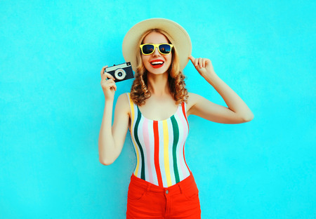Colorful happy smiling young woman holding retro camera in summer straw hat having fun on blue wall...