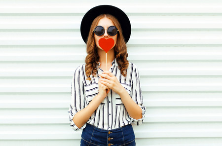 Portrait young woman kissing red heart shaped lollipop or hides her lips in black round hat, white striped shirt on white wall backgroundの写真素材
