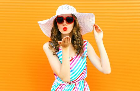 Portrait pretty woman blowing red lips sends sweet air kiss wearing summer straw hat, colorful striped dress on orange wall backgroundの写真素材