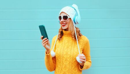 Winter portrait happy smiling woman holding phone in wireless headphones listening to music wearing...