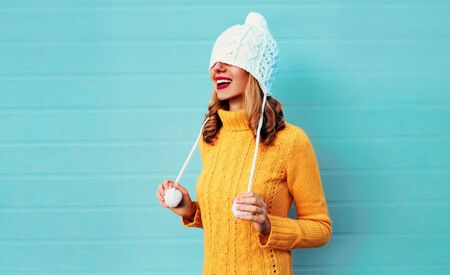 Winter portrait happy smiling young woman having fun pulls a hat over her eyes wearing yellow knitted sweater and white hat with pom pom on blue wall backgroundの写真素材