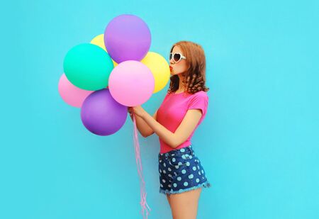 Lovely young woman holding in hands and kissing bunch of balloons wearing a shorts and pink t-shirt on blue wall background, side viewの写真素材