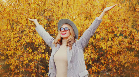 Autumn day, stylish young woman raising her hands up wearing gray coat, round hat over yellow leaves...