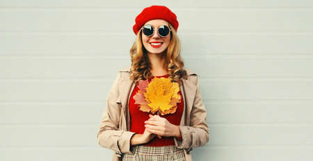 Autumn portrait of smiling young woman with yellow maple leaves wearing red french beret over gray backgroundの写真素材