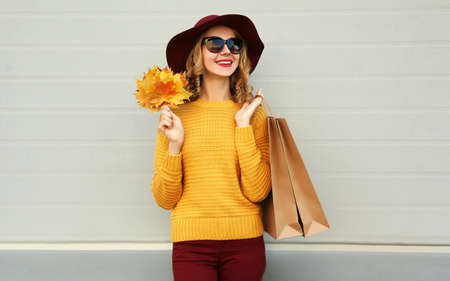 Autumn portrait of woman with shopping bags and yellow maple leaves wearing a sweater, hat over gray...
