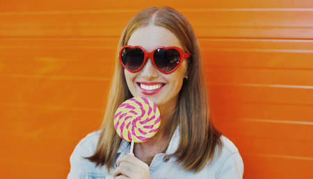 portrait close up of happy smiling woman with lollipop in red heart shaped sunglasses over an orange...