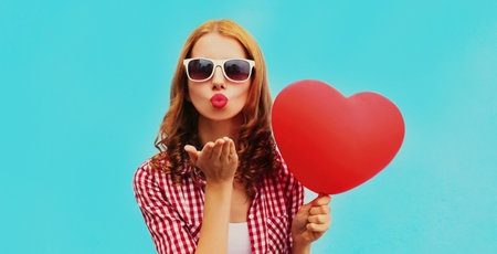 Portrait of happy young woman holding red heart shaped balloon and blowing lips sending sweet air kiss on a blue backgroundの写真素材