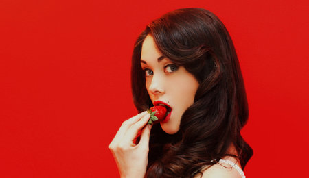 Portrait close up of attractive brunette sexy woman enjoying a taste of fresh strawberry on a colorful red backgroundの写真素材