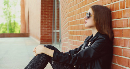 Young woman in casual sitting in the city wearing a black rock style over a brick wall backgroundの写真素材
