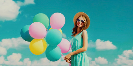 Happy smiling young woman with bunch of colorful balloons wearing a summer straw hat on the field on a blue sky backgroundの写真素材