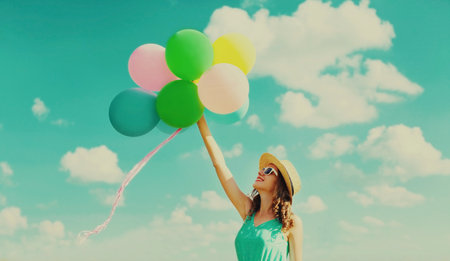 Happy smiling young woman with bunch of colorful balloons wearing a summer straw hat on the field on a blue sky backgroundの写真素材