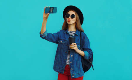 Portrait of stylish woman taking selfie picture by smartphone wearing a black hat, backpack on a blue backgroundの写真素材