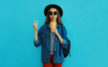 Portrait of young woman drinking a juice wearing a black round hat, denim jacket on a blue backgroundの写真素材