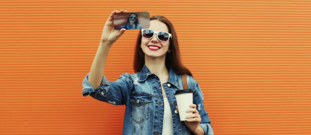 Portrait of smiling woman taking a selfie picture by smartphone wearing a denim jacket on a orange...