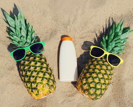 Close up of summer two funny pineapple in colorful sunglasses and sunscreen skin bottle on a beachの写真素材