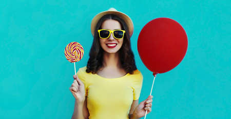 Portrait of happy smiling woman with lollipop and red balloon wearing a summer straw hat on a blue backgroundの写真素材