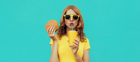 Portrait close up of young woman with fast food, burger and cup of juice wearing a yellow t-shirt, sunglasses on a blue backgroundの写真素材