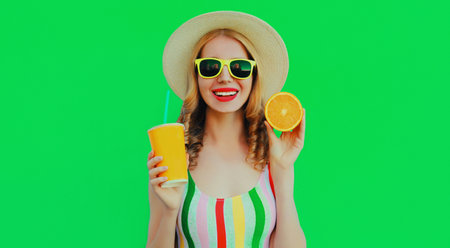 Summer portrait of happy smiling young woman with fresh cup of juice and slice of orange fruit wearing a straw hat, sunglasses on green backgroundの写真素材