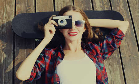 Portrait of happy smiling young woman photographer with vintage film camera lying on a skateboard outdoorsの写真素材