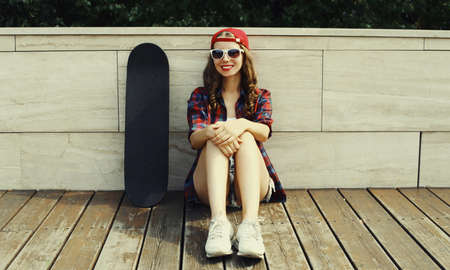Happy smiling young woman sitting with skateboard wearing a baseball cap on city streetの写真素材