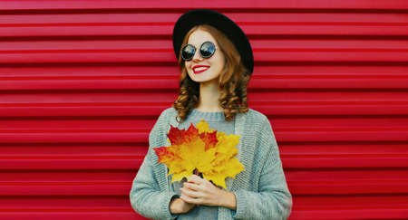 Autumn portrait of happy smiling young woman with yellow maple leaves wearing a black round hat,...