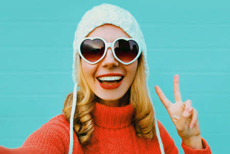 Winter portrait of happy smiling young woman stretching hand for taking selfie wearing a red knitted sweater, white hat, heart shaped sunglasses on blue wall backgroundの写真素材