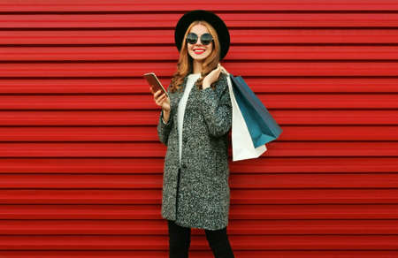 Portrait of happy smiling woman with shopping bags and smartphone wearing a gray coat, hat on colorful red background