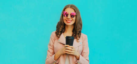 Portrait of beautiful smiling young woman with cup of coffee wearing a leather jacket on blue...