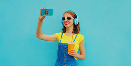 Portrait of happy smiling young woman taking a selfie by smartphone in headphones listening to music on blue backgroundの写真素材
