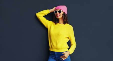 Portrait of stylish smiling young woman wearing an yellow knitted sweater and pink hat on black wall...