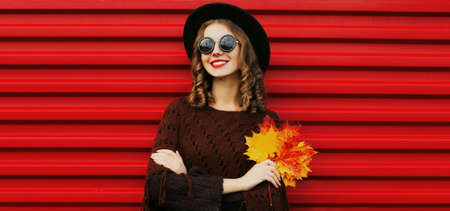 Portrait of beautiful young woman with yellow maple leaves wearing a brown knitted poncho, black...