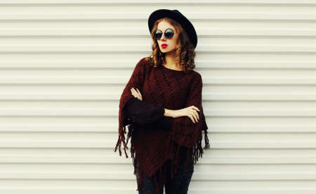 Portrait of beautiful young woman wearing a brown knitted poncho, black round hat on white...