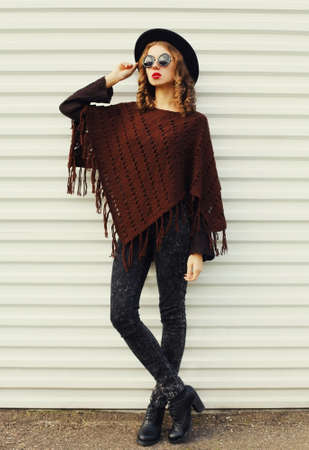 Portrait of beautiful young woman wearing a brown knitted poncho, black round hat on white...